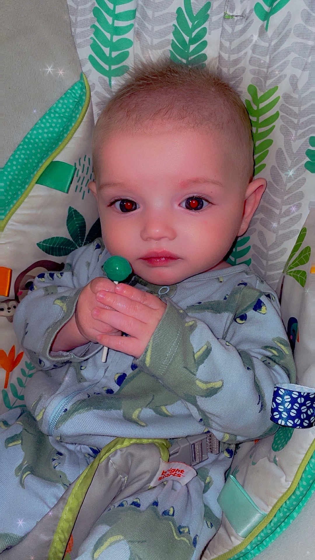 Sylas joined the competition — help win amazing prizes! baby, child, lollipop, onesie, dinosaur_pattern, seat, patterned_fabric, hand, face, red_eye, indoor, cute, infant, clothing, baby_seat, holding, sparkle_effect, expression, young_child, cozy