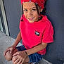 black_shoes, braided_hair, casual_clothing, child, concrete, crouching, cute, daylight, denim_shorts, fashion, happy, outdoor, person, portrait, red_bandana, red_shirt, smiling, sneakers, wall, young