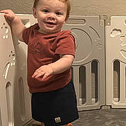 Morgan is registered to the contest to win money with this photo: baby, barefoot, carpet, child, curly_hair, floor, happy, indoor, person, play_area, playpen, red_hair, shorts, smiling, standing, toddler, toy, tshirt, wall, young_child