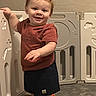 baby, barefoot, carpet, child, curly_hair, floor, happy, indoor, person, play_area, playpen, red_hair, shorts, smiling, standing, toddler, toy, tshirt, wall, young_child