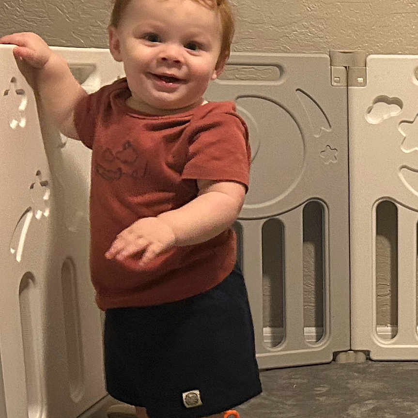 Morgan is registered to the contest to win money with this photo: baby, barefoot, carpet, child, curly_hair, floor, happy, indoor, person, play_area, playpen, red_hair, shorts, smiling, standing, toddler, toy, tshirt, wall, young_child