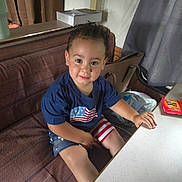Zyalle is registered to the contest to win money with this photo: toddler, child, smile, blue_shirt, american_flag, shorts, striped_shorts, couch, table, purse, curtains, box_of_crayons, indoor, person, sitting, happy, furniture, face, home, play