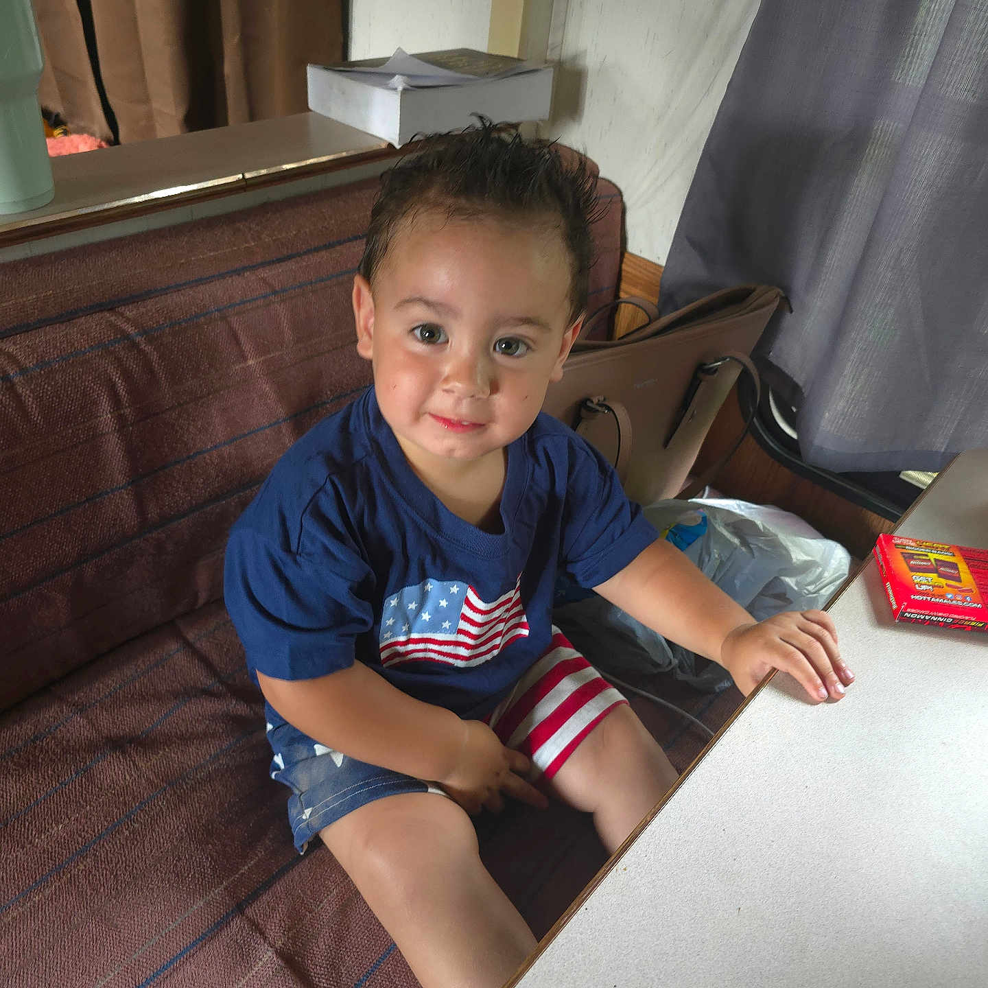 Zyalle is registered to the contest to win money with this photo: toddler, child, smile, blue_shirt, american_flag, shorts, striped_shorts, couch, table, purse, curtains, box_of_crayons, indoor, person, sitting, happy, furniture, face, home, play