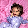 baby, child, onesie, purple_clothing, heart_pattern, pink_blanket, smiling, happy, cute, face, hand, blue_eyes, curly_hair, black_hair_bow, infant, portrait, soft_texture, lying_down, indoors, toddler