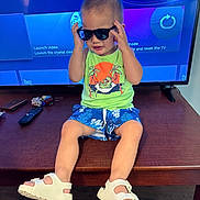 Alexander is registered to the contest to win money with this photo: child, toddler, portrait, sunglasses, smiling, sitting, television, indoors, living_room, green_shirt, blue_shorts, sandals, remote_control, furniture, wooden_table, floor, electronics, fashion, playful, casual_clothing