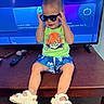 child, toddler, portrait, sunglasses, smiling, sitting, television, indoors, living_room, green_shirt, blue_shorts, sandals, remote_control, furniture, wooden_table, floor, electronics, fashion, playful, casual_clothing