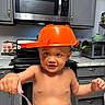 baby, child, toddler, kitchen, cooking, orange_hat, pan, fork, spoon, salad, vegetables, lettuce, bowl, countertop, microwave, cabinets, appliance, shirtless, portrait, indoors