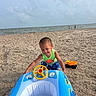 Alexander joined the competition — help win amazing prizes! child, toddler, baby, beach, ocean, sea, sand, inflatable_boat, float, toy_truck, steering_wheel, swimwear, green_shirt, blue_shorts, outdoor, vacation, shoreline, cloudy_sky, sitting, playtime