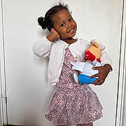 Keyah participe au concours pour gagner de l'argent avec cette photo : child, girl, toy, doll, indoor, smiling, cute, dress, cardigan, pink, patterned_clothing, curly_hair, hair_buns, standing, floor, door, wooden_floor, playful, happy, portrait