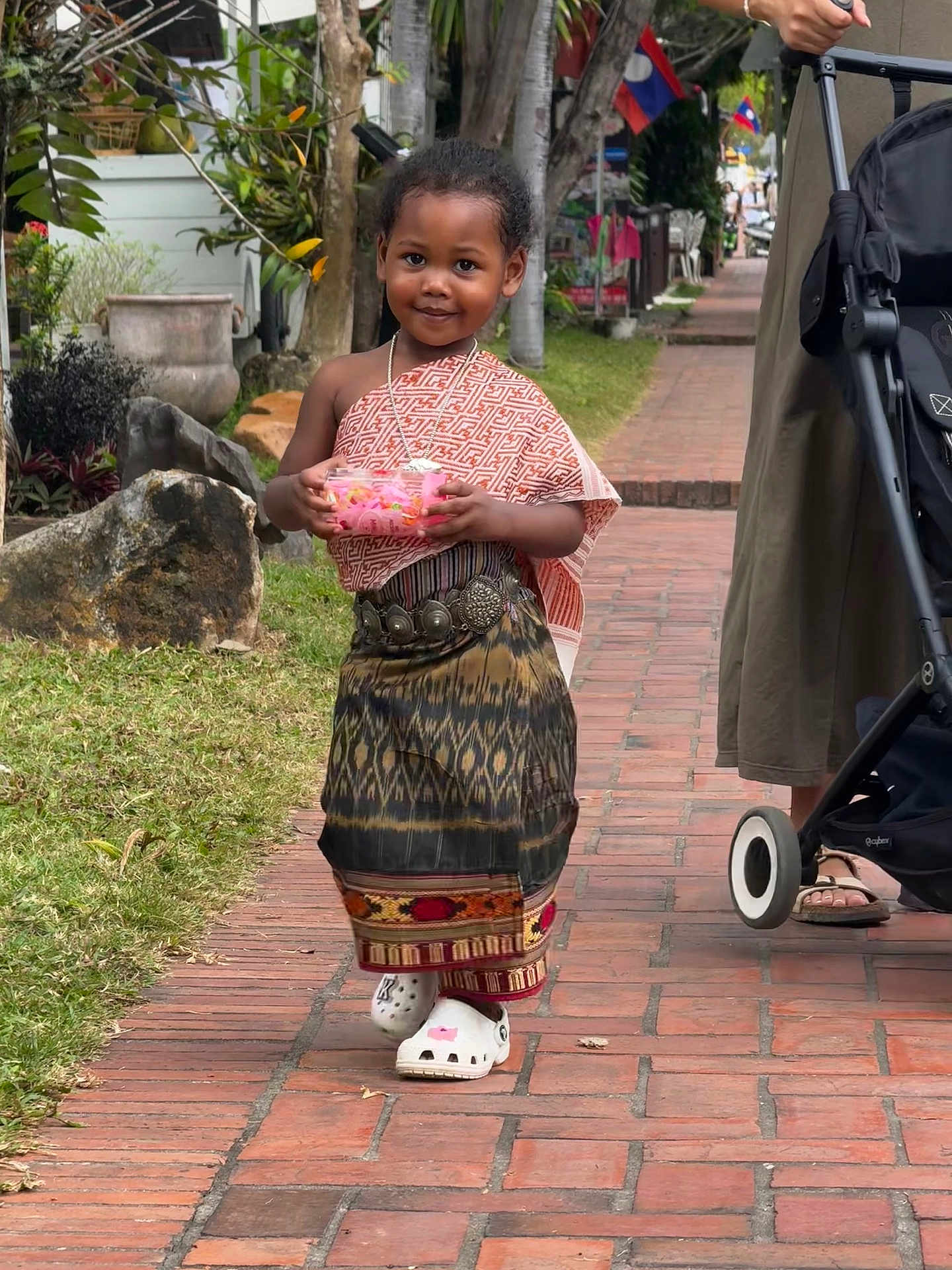 Keyah participe au concours pour gagner de l'argent avec cette photo : child, traditional_clothing, walking, outdoor, brick_pathway, stroller, adult, greenery, plants, trees, flags, smiling, person, footwear, sidewalk, daylight, necklace, container, street, public_place