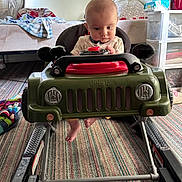 Weston is registered to the contest to win money with this photo: baby, baby_walker, bed, blanket, car_toy, child, clothing, curious, floor, foot, furniture, head, indoor, jeep, plastic, play, room, sitting, striped_carpet, toy