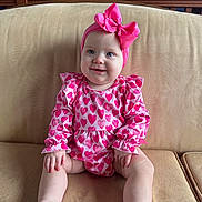 Raleigh is registered to the contest to win money with this photo: baby, infant, headband, bow, pink, hearts_pattern, onesie, couch, sofa, sitting, smile, chubby_cheeks, portrait, indoor, legs, feet, hands, blue_eyes, furniture, adorable
