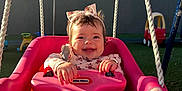 Fernanda is registered to the contest to win money with this photo: baby, child, swing, pink, smiling, headband, outdoor, playground, toy_car, grass, sunlight, happy, person, feet, clothing, rope, seat, infant, cute, playtime