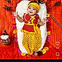 baby, child, traditional_clothing, yellow, red, peacock_feathers, flowers, orange_background, smiling, cushion, festive, lying_down, headband, bracelet, portrait, indian_culture, celebration, decor, happy, infant