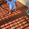 toddler, child, outdoor, deck, wooden_floor, sunlight, shadow, blue_outfit, sandals, person, daylight, fence_shadow, smiling, walking, young_child, playful, summer, casual_clothing, happy, exploring