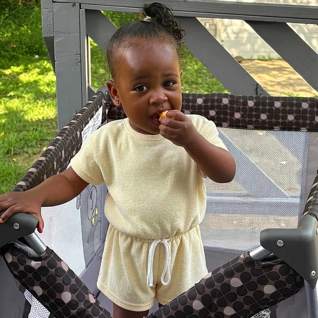 Alicia Goldsby is registered to the contest to win money with this photo: child, toddler, playpen, snack, outdoor, greenery, sunlight, curly_hair, shorts, top, standing, fence, daylight, backyard, person, baby, summer_clothing, cute, leisure, play
