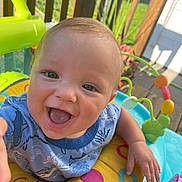 Mason is registered to the contest to win money with this photo: baby, child, smiling, happy, play_center, colorful, outdoor, sunlight, toy, infant, face, hand, clothing, blue_shirt, yellow_ring, green_toy, wooden_deck, fence, flower_pot, portrait