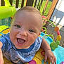 baby, child, smiling, happy, play_center, colorful, outdoor, sunlight, toy, infant, face, hand, clothing, blue_shirt, yellow_ring, green_toy, wooden_deck, fence, flower_pot, portrait