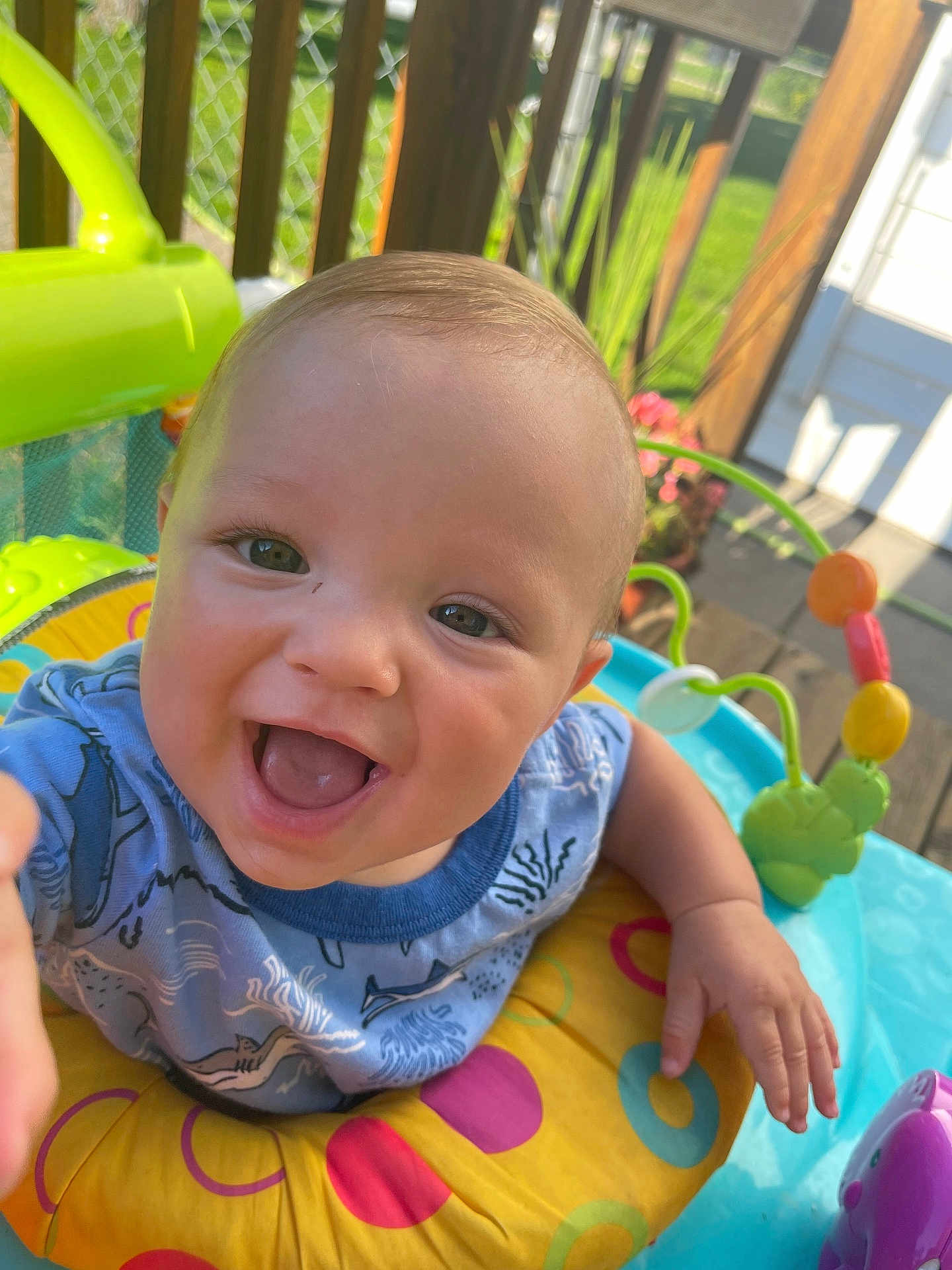Mason is registered to the contest to win money with this photo: baby, child, smiling, happy, play_center, colorful, outdoor, sunlight, toy, infant, face, hand, clothing, blue_shirt, yellow_ring, green_toy, wooden_deck, fence, flower_pot, portrait