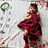 baby, infant, red_dress, headband, blanket, floral_pattern, milestone_tracker, numbers, lying_down, cute, smiling, child, soft_texture, indoors, portrait, baby_boots, month_marker, colorful, celebration, happy