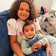 Kaden is registered to the contest to win money with this photo: child, baby, smiling, couch, curly_hair, confetti, overalls, plaid_shirt, bracelet, pillow, living_room, window_blind, cap, happy, person, portrait, seated, family, indoor, cute