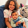 child, baby, smiling, couch, curly_hair, confetti, overalls, plaid_shirt, bracelet, pillow, living_room, window_blind, cap, happy, person, portrait, seated, family, indoor, cute