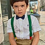 bow_tie, boy, brown_hair, casual, child, expression, face, fashion, outdoor, pants, person, portrait, serious, shirt, short_sleeve, sitting, street, suspenders, window_reflection, young