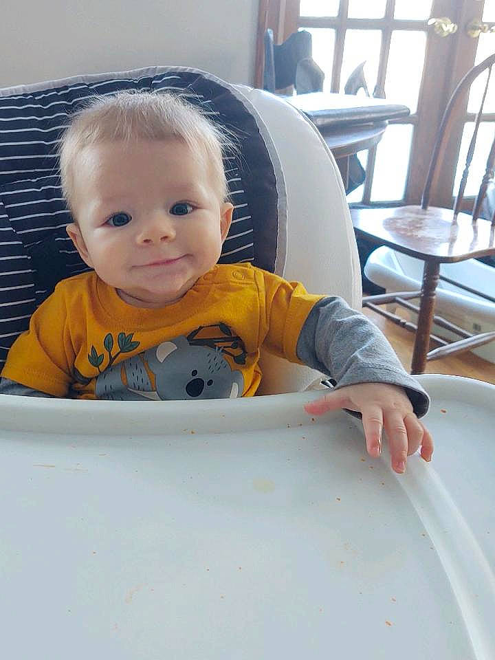 Fletcher is registered to the contest to win money with this photo: baby, baby_toddler_clothing, bib, chair, cheek, child, comfort, flooring, fun, happy, person, product, room, sitting, sleeve, smile, t_shirt, table, toddler, vacation