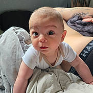 Finn is registered to the contest to win money with this photo: baby, infant, blanket, bed, tattoo, arm, person, indoor, curious, wide_eyes, white_onesie, wooden_chair, soft_lighting, skin, expression, candid, closeup, human, home, resting