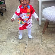 Nyla joined the competition — help win amazing prizes! toddler, child, smiling, laughing, indoor, floor, tile, dress, clothing, flower_lei, happy, person, cute, standing, white_socks, hair_bow, joyful, home, furniture, chair