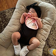 Braelynn joined the competition — help win amazing prizes! baby, pacifier, headband, bib, sneakers, black_outfit, cushion, wooden_floor, carpet, baby_bottle, infant, child, indoor, cute, relaxed, soft_lighting, portrait, closeup, homedecor, comfort