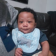 Takkari joined the competition — help win amazing prizes! baby, infant, child, portrait, smiling, sitting, couch, indoors, home, bib, elephant, stars, blue_clothing, curly_hair, brown_skin, face, hands, arm, furniture, family