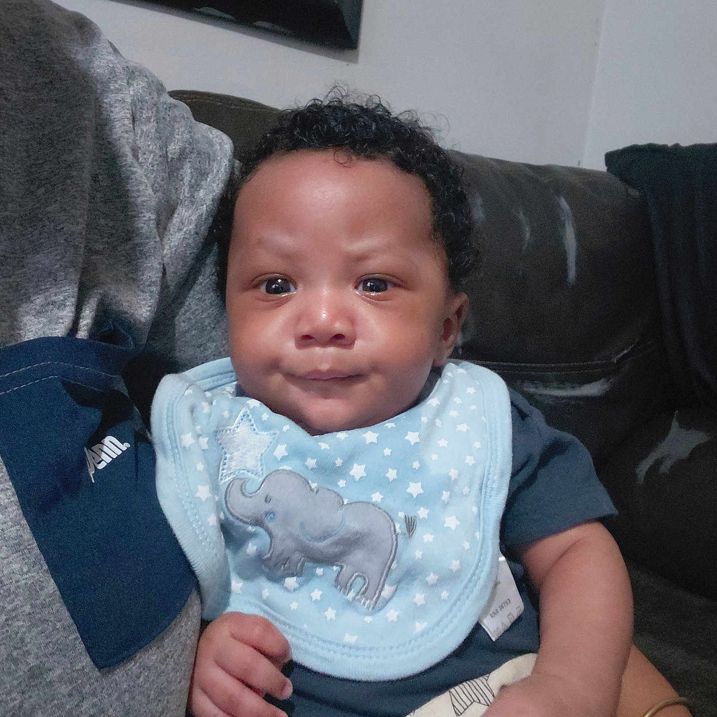 Takkari joined the competition — help win amazing prizes! baby, infant, child, portrait, smiling, sitting, couch, indoors, home, bib, elephant, stars, blue_clothing, curly_hair, brown_skin, face, hands, arm, furniture, family