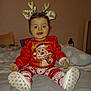 baby, child, smiling, reindeer_headband, holiday_clothing, red_sweater, patterned_pants, bed, pillow, blanket, indoors, person, cute, festive, christmas, happy, sitting, footwear, cozy, portrait