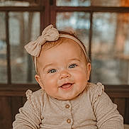 Daisy is registered to the contest to win money with this photo: baby, smiling, headband, bow, infant, portrait, face, cute, child, window, wood, warm_lighting, indoor, clothing, happy, skin, eyes, head, sitting, young