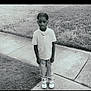 child, sidewalk, grass, outdoor, polo_shirt, pants, shoes, beaded_hair, standing, monochrome, portrait, casual_clothing, young, person, alone, quiet, daytime, urban, background, calm