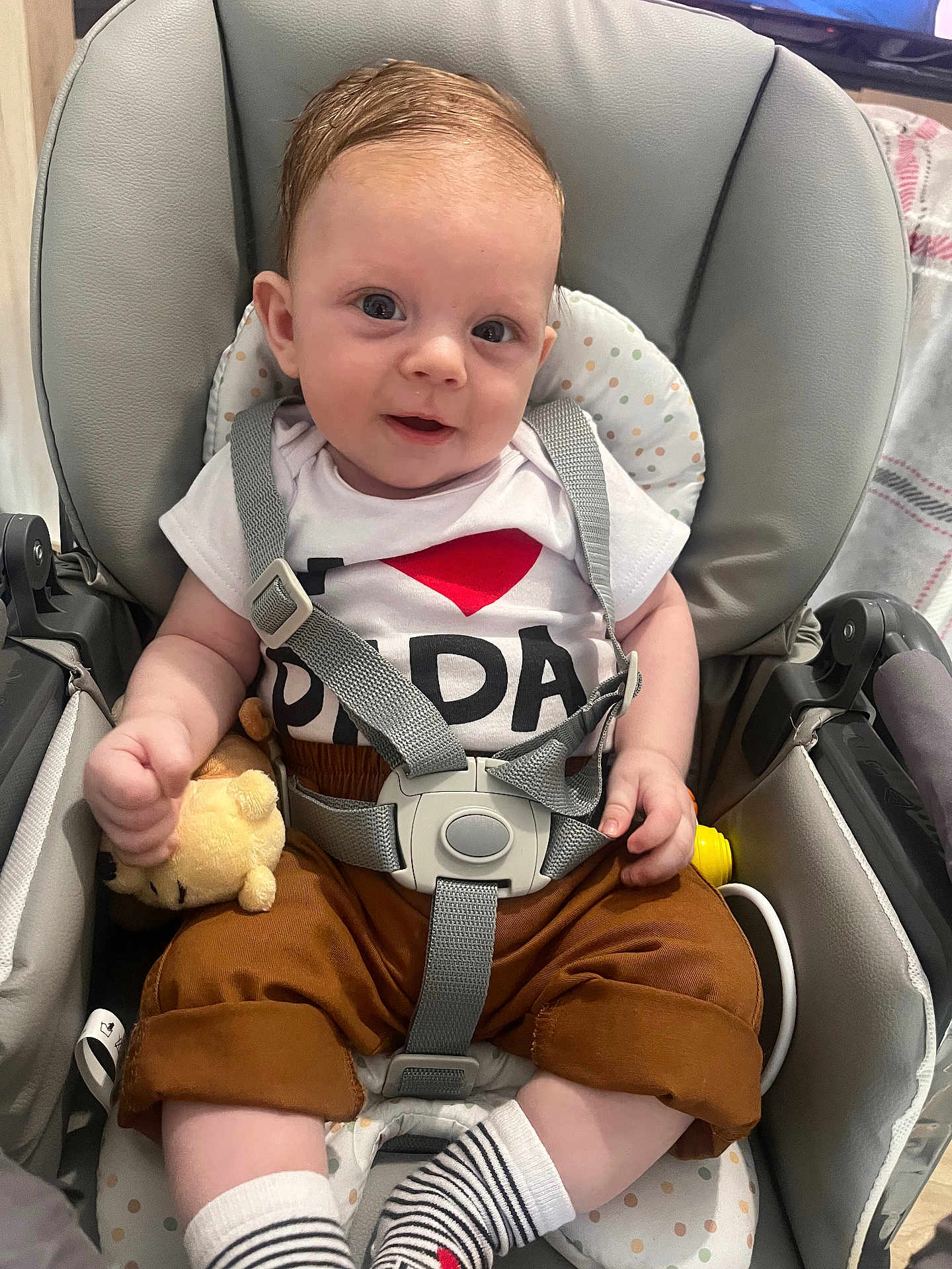 Tommy a rejoint le concours — aidez-le/la à gagner de superbes lots ! baby, child, car_seat, seatbelt, plush_toy, white_shirt, brown_pants, striped_socks, smiling, indoors, cute, infant, person, happy, toddler, clothing, seat, baby_toy, safety_harness, portrait