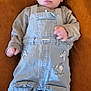 baby, child, infant, overalls, cap, lying_down, brown_background, plush_toy, grey_shirt, cute, tongue_out, warm_clothing, small_hands, face, headwear, footwear, pocket, closeup, indoor, soft_texture