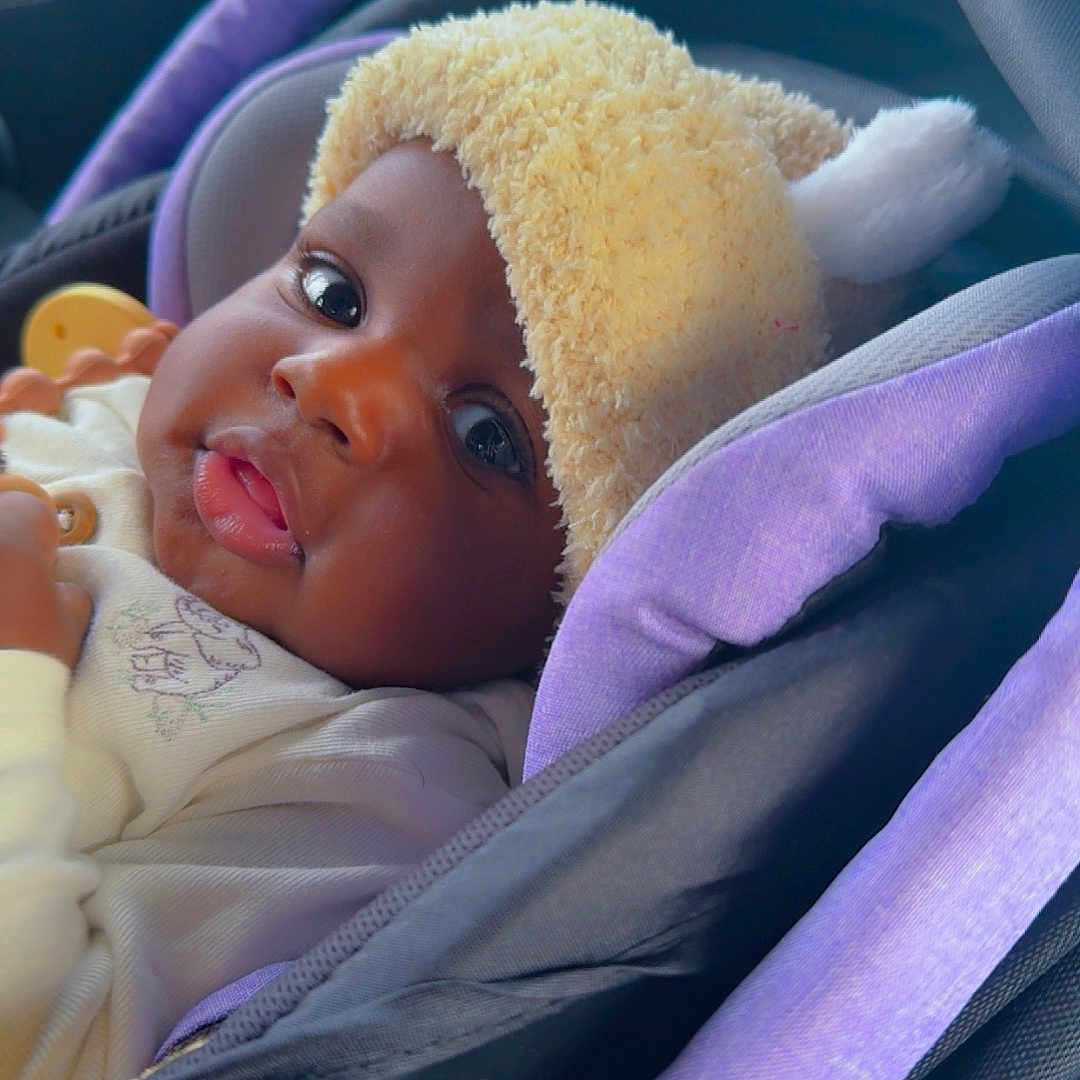 Abdoulaye Ba is registered to the contest to win money with this photo: baby, black, car, car_seat, child, closeup, clothing, cozy, cute, eyes, face, fuzzy_hat, hat, indoors, infant, newborn, portrait, purple, seatbelt, young_child
