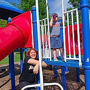 Hailey is registered to the contest to win money with this photo: child, playground, play_structure, slide, bottle, person, red_hair, blue, red, green_trees, sunny, outdoor, grass, smile, casual_clothing, fence, wood_chips, daytime, happy, park
