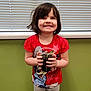 Olivia is registered to the contest to win money with this photo: child, smiling, indoor, plant, soil, cup, holding, short_hair, red_shirt, cartoon_shirt, gray_pants, window_blinds, green_wall, standing, happy, young, person, casual_clothing, floor, carpet