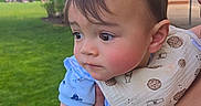 Aiden is registered to the contest to win money with this photo: adult_arm, baby, bib, blue_clothing, child, curious, daylight, face, grass, green, holding, nature, outdoor, park, person, portrait, skin, sunny, tree, young_child