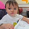Aiden joined the competition — help win amazing prizes! toddler, child, high_chair, white_shirt, indoor, restaurant, table, hand, face, curious, colorful, wall_art, orange, booth_seating, expression, young_child, portrait, casual, seated, cute
