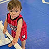 toddler, child, red_jersey, sportswear, barefoot, indoor, blue_floor, plastic_bar, curious, young_child, standing, play_area, gym_floor, person, baby, active, hands, hair, expression, concentration