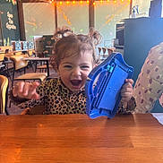 Inaya a rejoint le concours — aidez-le/la à gagner de superbes lots ! background, blue_toy, cafe, celebration, chair, child, furniture, happy, indoor, leopard_print_clothing, person, pigtails, playful, smiling, string_lights, table, toddler, toy, window, wooden_table