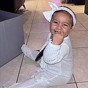 Inaya participe au concours pour gagner de l'argent avec cette photo : baby, toddler, smiling, sitting, hand_in_mouth, headband, bow, onesie, footed_pajamas, white_clothing, tile_floor, indoors, living_room, storage_box, remote_control, happy, cheeks, cute, portrait, infant