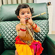 Veda is registered to the contest to win money with this photo: baby, beads, bracelets, child, colorful, cultural, curly_hair, cute, decor, expression, flute, green_chair, indoor, jewelry, person, playing, portrait, sitting, toddler, traditional_clothing