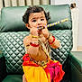baby, beads, bracelets, child, colorful, cultural, curly_hair, cute, decor, expression, flute, green_chair, indoor, jewelry, person, playing, portrait, sitting, toddler, traditional_clothing