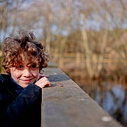 Marley joined the competition — help win amazing prizes! child, curly_hair, smile, outdoor, bridge, wooden_railing, nature, trees, water, winter_coat, fur_hood, portrait, young, person, cold_weather, daylight, happy, playful, park, forest
