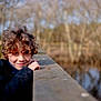 child, curly_hair, smile, outdoor, bridge, wooden_railing, nature, trees, water, winter_coat, fur_hood, portrait, young, person, cold_weather, daylight, happy, playful, park, forest